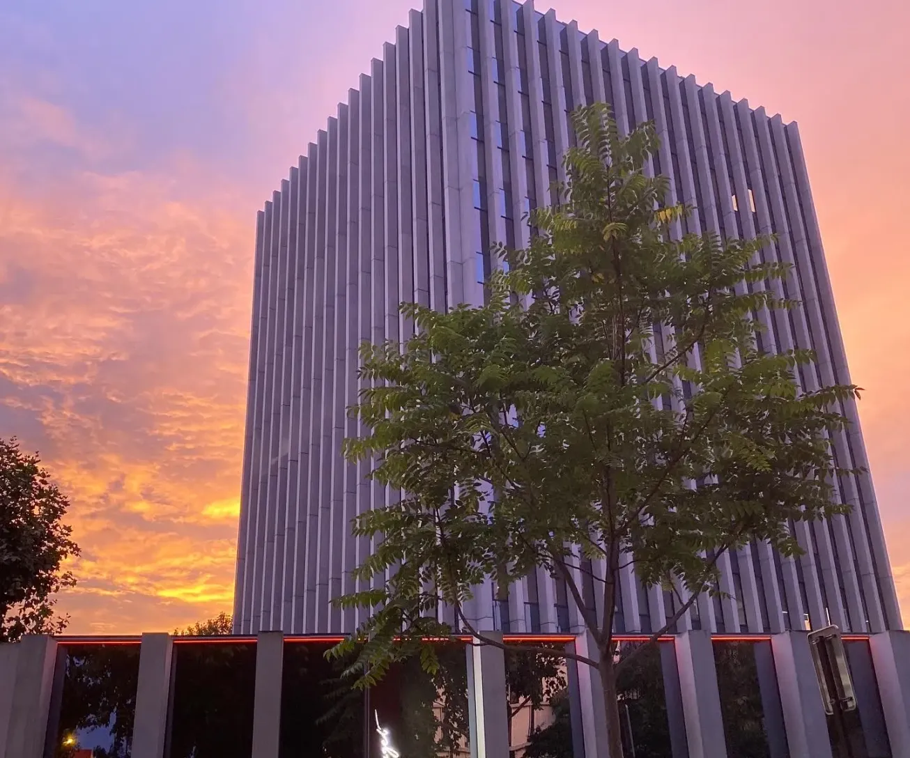 Bâtiment moderne à façade verticale striée avec des arbres devant, sous un ciel de coucher de soleil aux teintes orangées et roses