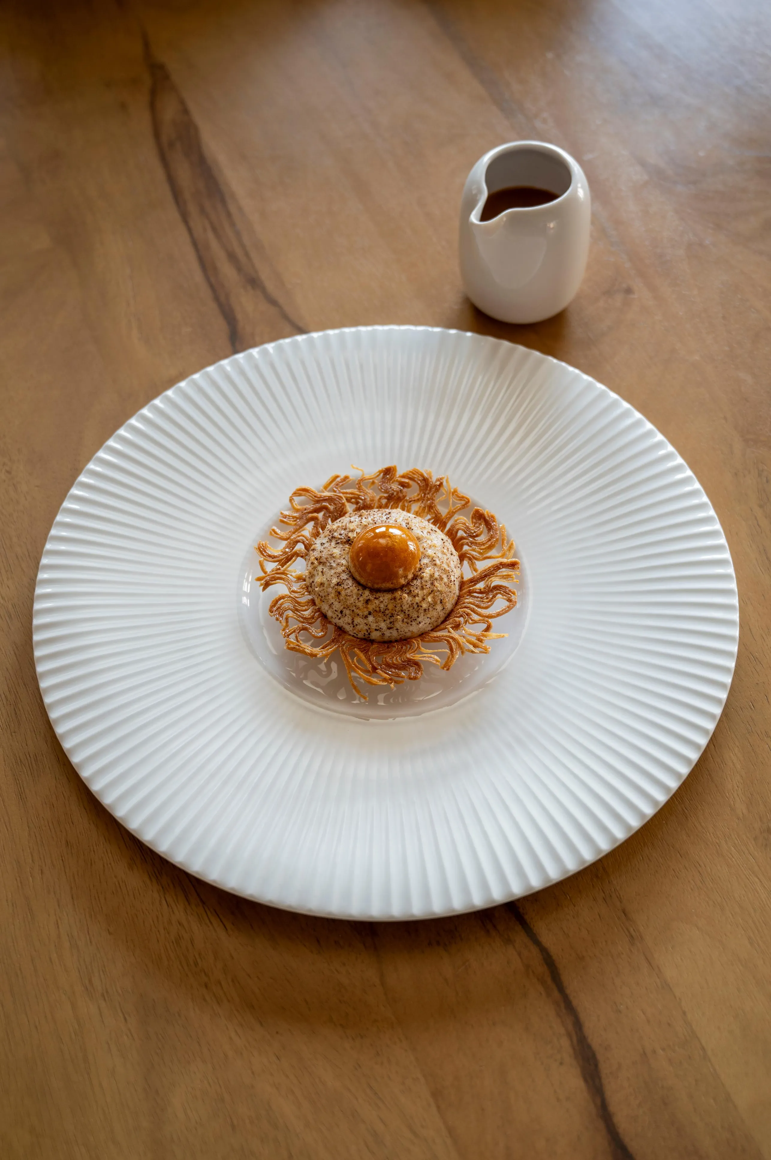 Un dessert composé d'une boule de glace saupoudrée de cacao et entouré de vermicelles dorés, servi sur une assiette blanche avec une petite verseuse à côté